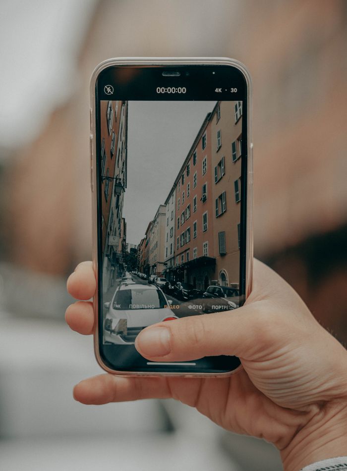 A hand holding a smartphone capturing an urban street view with buildings and cars.